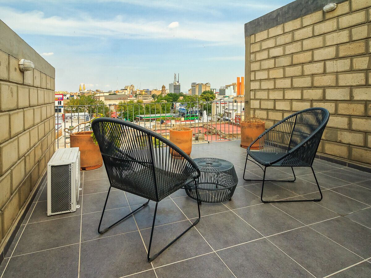 Una terraza en la azotea con dos sillas de malla metálica negra y una pequeña mesa redonda, una planta en maceta y un radiador.