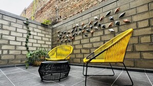 Eine Ziegelwand mit verzierten Metallspitzen, zwei gelbe Stühle und ein schwarzer Flechtstuhl auf einer gefliesten Terrasse.