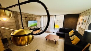 Ein modernes Wohnzimmer mit einem großen Metalltopf, einer schwarzen Couch mit gelben Kissen, einem Holztisch, einem Fernseher an der Wand und Blick durch große Fenster.