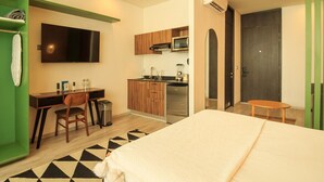 A hotel room with a bed, a desk, a chair, a TV, a kitchenette, and a green wardrobe.