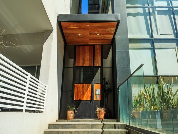 Una entrada moderna de edificio con puerta de madera, barandilla de vidrio y plantas en macetas.