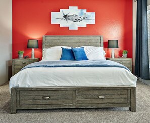 Een bed met houten frame, witte beddengoed en blauwe kussens. Twee nachtkastjes met lampen en kleine planten staan ernaast. Achter het bed een rode muur met een lijst eromheen.