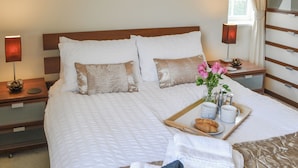 Un lit parfaitement fait, avec du linge de lit blanc, un plateau avec des croissants et des tasses de café, et un vase rempli de fleurs roses.