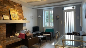 A living room with a brick fireplace, a television, a glass dining table, and a large window. - Charming sea house (Saint-Valery-en-Caux)