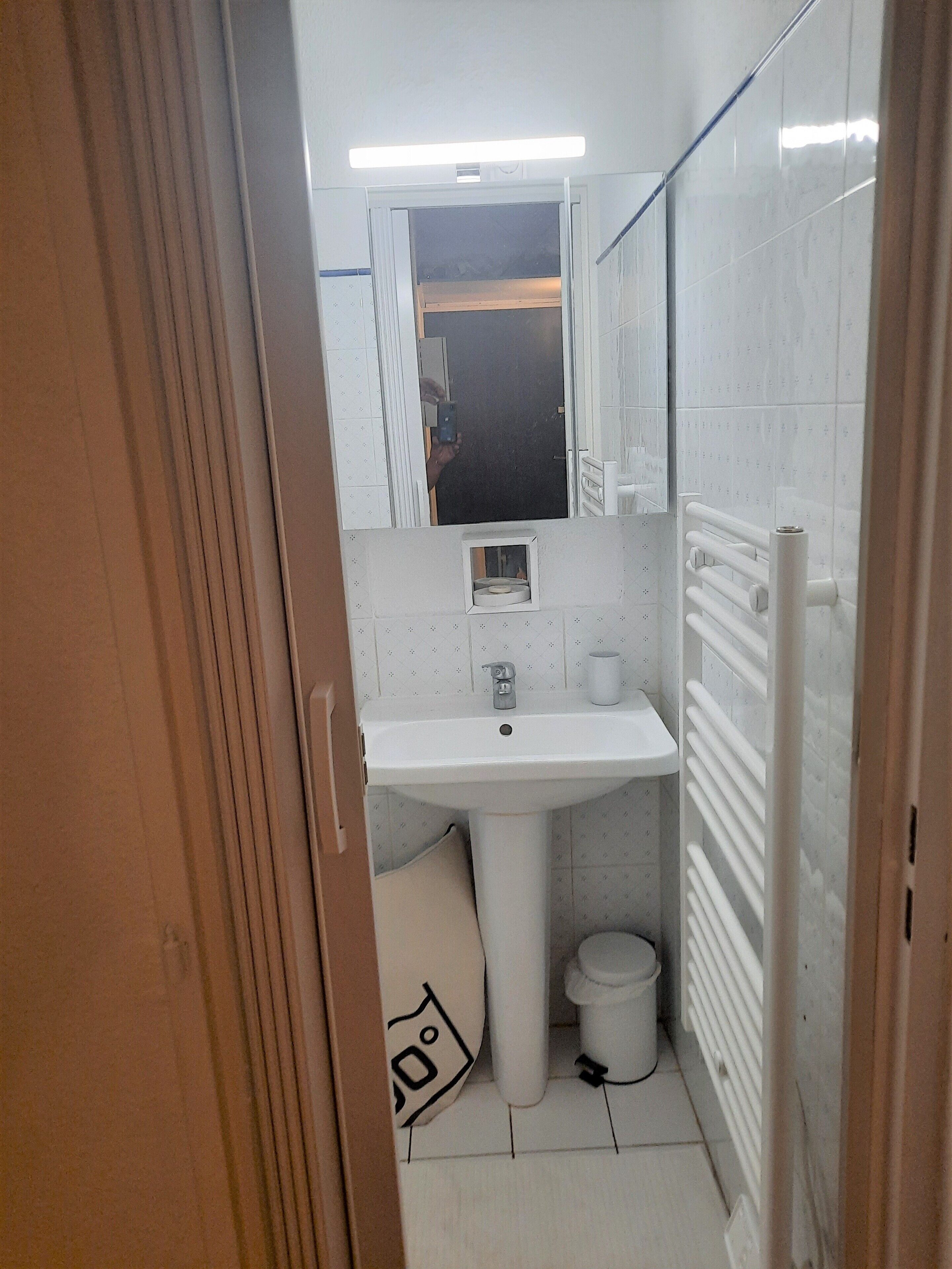 A small, compact bathroom with a white sink, mirror, and toilet. There is a towel rack and a small trash bin.