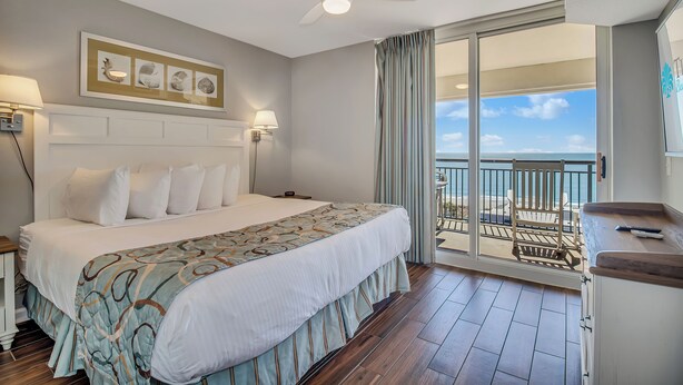 Habitación de hotel con una cama grande, ventilador de techo, balcón con vista al océano y escritorio con silla.