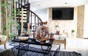 A modern living room with a curved staircase, a flat-screen TV, a dining table with wine glasses, and a potted plant.