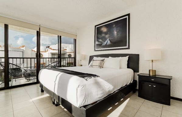A modern hotel room with a large bed, a nightstand, a framed picture of a night sky, and a balcony with a view of buildings.