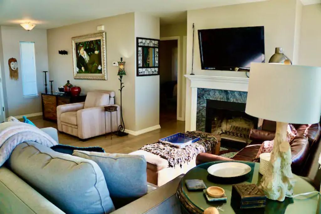 Una sala de estar con un sofá, un sillón y una chimenea. Hay un televisor montado sobre la chimenea y un cuadro en la pared.