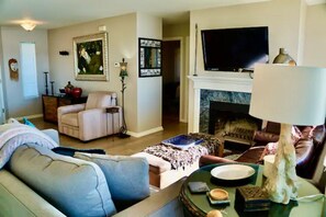Een woonkamer met een bank, fauteuil en een open haard. Een televisie is boven de open haard bevestigd en er hangt een lijst met een afbeelding aan de muur.