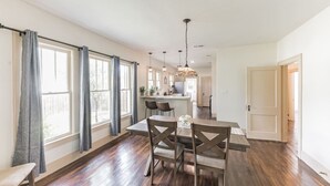 A spacious dining area with a wooden table and chairs, large windows with curtains, a kitchen area in the background, and a hanging light fixture. - Charming, Tudor Style Home Newly Renovated! (Mineral Wells)