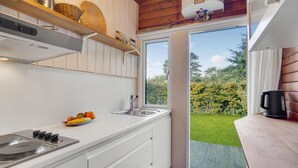 Une petite cuisine équipée d’un évier, d’une cuisinière et d’un saladier rempli de fruits. Une fenêtre donne sur une cour verdoyante.