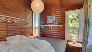 Une chambre dans une cabane en bois, avec un lit, un luminaire suspendu, un tableau et une fenêtre donnant sur la forêt.
