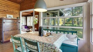 L’intérieur chaleureux d’un chalet, avec un revêtement mural en bois, une table à manger recouverte d’une nappe à motifs floraux, un banc agrémenté de coussins et une grande fenêtre offrant une vue sur les arbres.