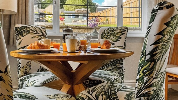 Et morgenbord med kager, kaffe og appelsinjuice, placeret i et rum med store vinduer der kigger ud over et træhegn og grønne planter.