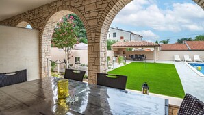 An outdoor patio with a stone archway, a dining table with chairs, a pool, and a lawn. - Beautiful apartment in Medulin with swimming pool (Medulin)