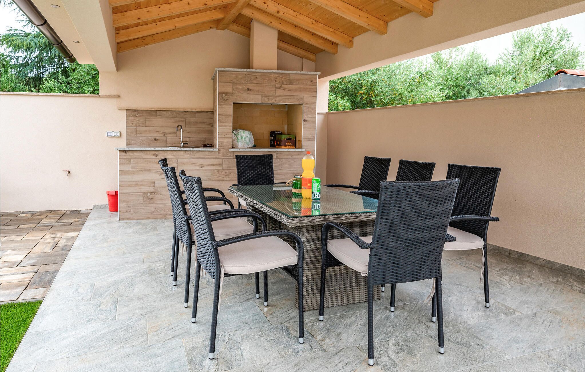 An outdoor patio with a glass-top table and six wicker chairs, a built-in grill, and a small sink.