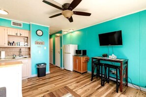A compact kitchen with wooden flooring, a ceiling fan, a microwave, a refrigerator, and a TV mounted on the wall.