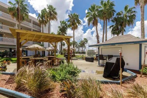 A covered outdoor area with tables and chairs, surrounded by palm trees and a building with balconies.