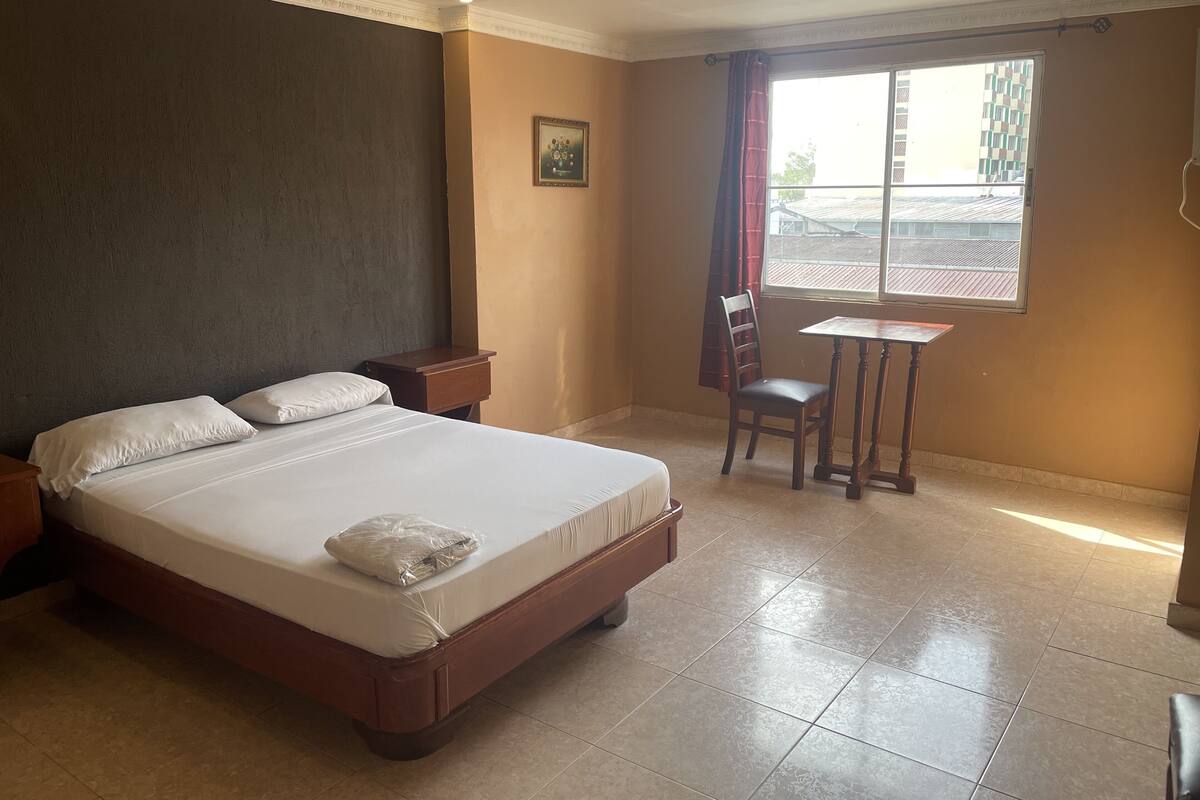 A hotel room with a large bed, a small table, a chair, a window with curtains, and a picture on the wall.