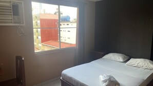 Una habitación de hotel con una cama, una silla, un ventanal con vistas a edificios y un equipo de aire acondicionado en la pared.