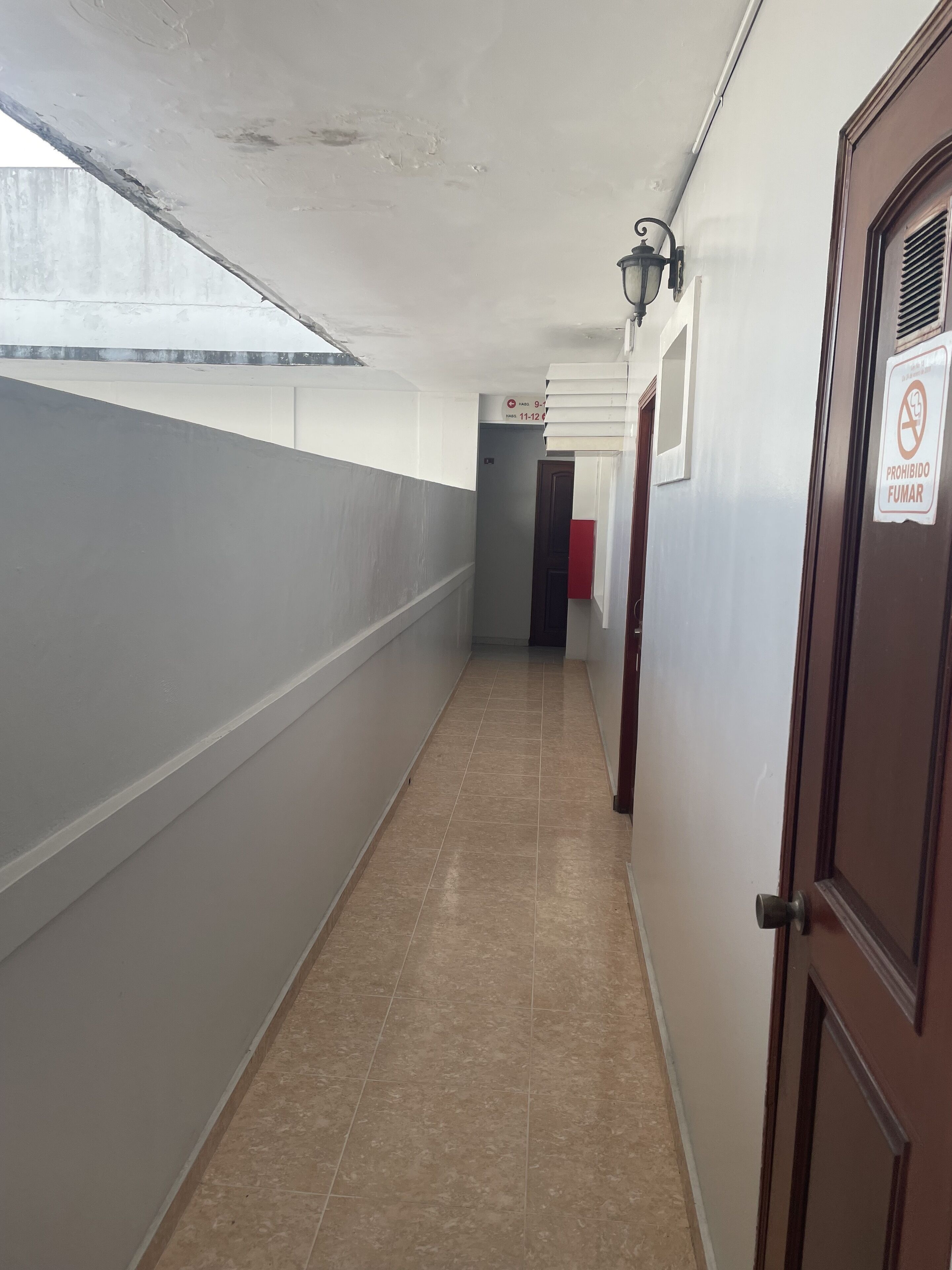 Un pasillo con suelo de baldosas, una puerta con un cartel de 'No Smoking' y una ventana a la izquierda.