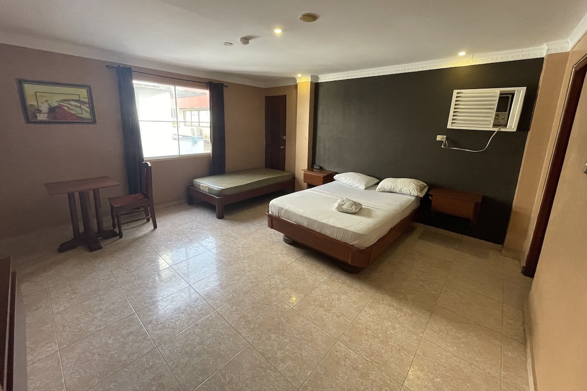 A bedroom with a bed, a small table, a bench, a window, and an air conditioning unit.