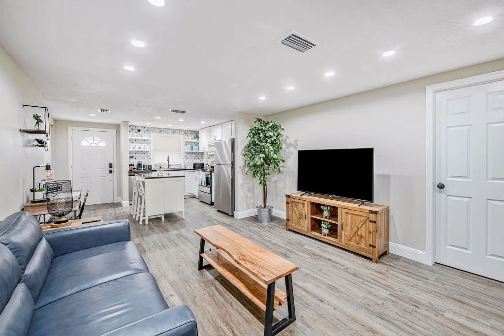 A modern living room with a leather sofa, a wooden coffee table, a TV on a wooden stand, and a kitchen in the background.