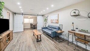 Ein modernes Wohnzimmer mit einer Couch, einem Couchtisch und einem Schreibtisch mit Stuhl. Links befindet sich ein Fernseher und im Hintergrund eine Essecke.
