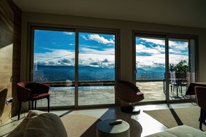 A room with a large sliding glass door leading to a balcony with a view of the sea and mountains. There are two red chairs and a round table with a remote control on it.