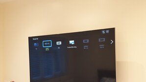 A flat-screen TV mounted on a wall with a source menu displaying various input options.
