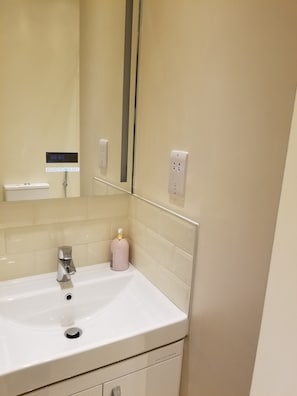 A bathroom with a sink, mirror, and shelf below, featuring a soap dispenser and a tiled wall with an electrical outlet.