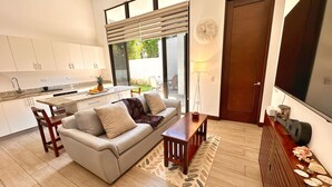 A modern living room with a sofa, coffee table, and a TV. There is a kitchen area with cabinets and a dining table. - Brand New! Jaco Beach outside the back gate! (Jaco)