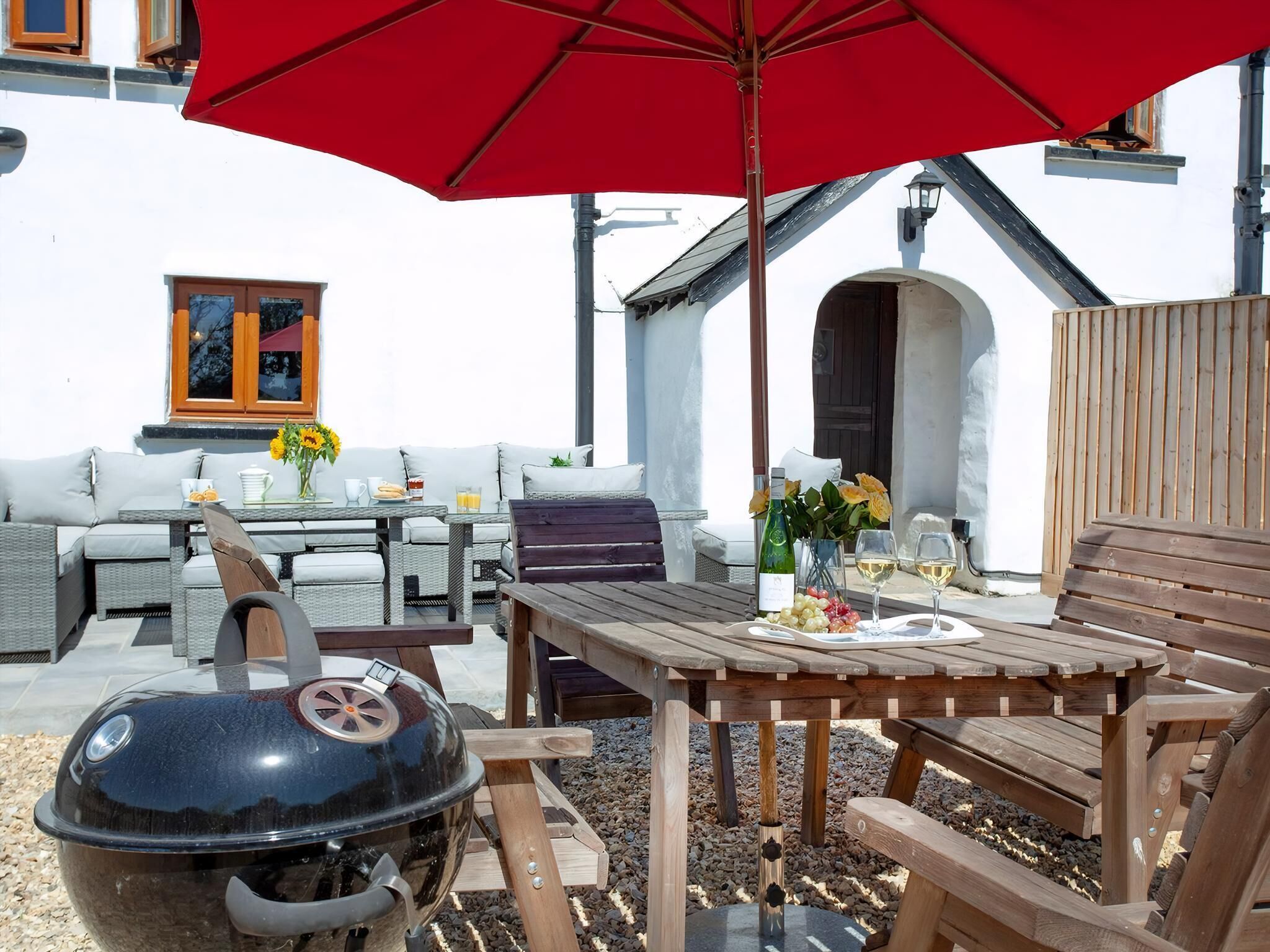 An outdoor patio with a red umbrella, wicker furniture, a wooden table, and a barbecue grill.