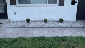 Uma casa com fachada branca, uma janela grande com persianas, um pequeno jardim de cascalho e dois arbustos, e uma porta de garagem preta.
