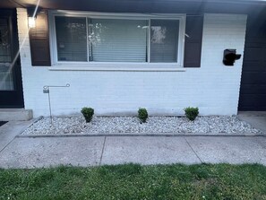 A house with a white exterior, a large window with blinds, a small garden with gravel and two shrubs, and a black garage door.