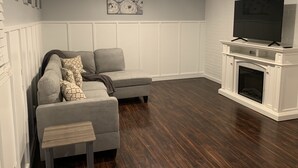Uma sala de estar com um sofá seccional cinza, uma mesinha de madeira, uma lareira e uma TV fixada na parede.
