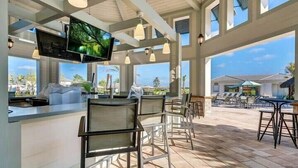 Un vaste espace extérieur aménagé pour les repas, avec de nombreuses tables et chaises, un coin bar doté d’un comptoir et de sièges, et de grandes fenêtres offrant une vue sur le paysage environnant.