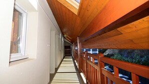 Un couloir aux poutres apparentes, éclairé par une lucarne, qui mène à un balcon doté d’une balustrade.