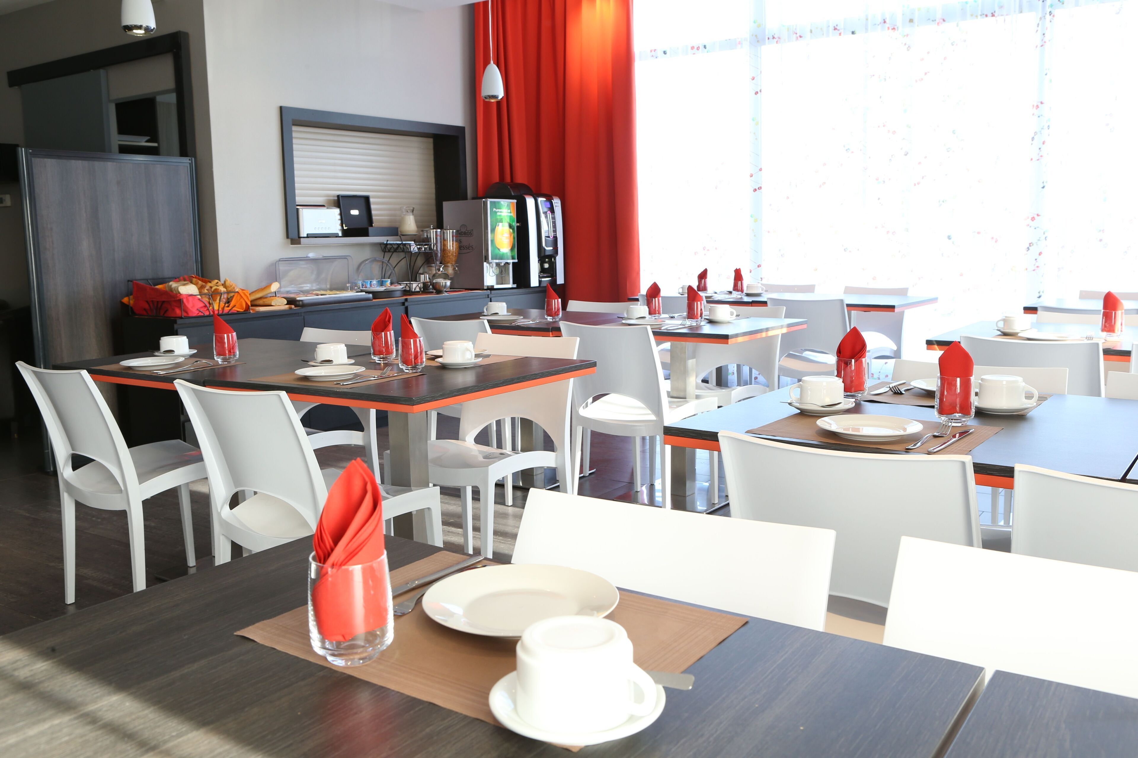 Un comedor moderno con sillas y mesas blancas, y servilletas rojas. Se observa una máquina de café y una encimera con un refrigerador al fondo.
