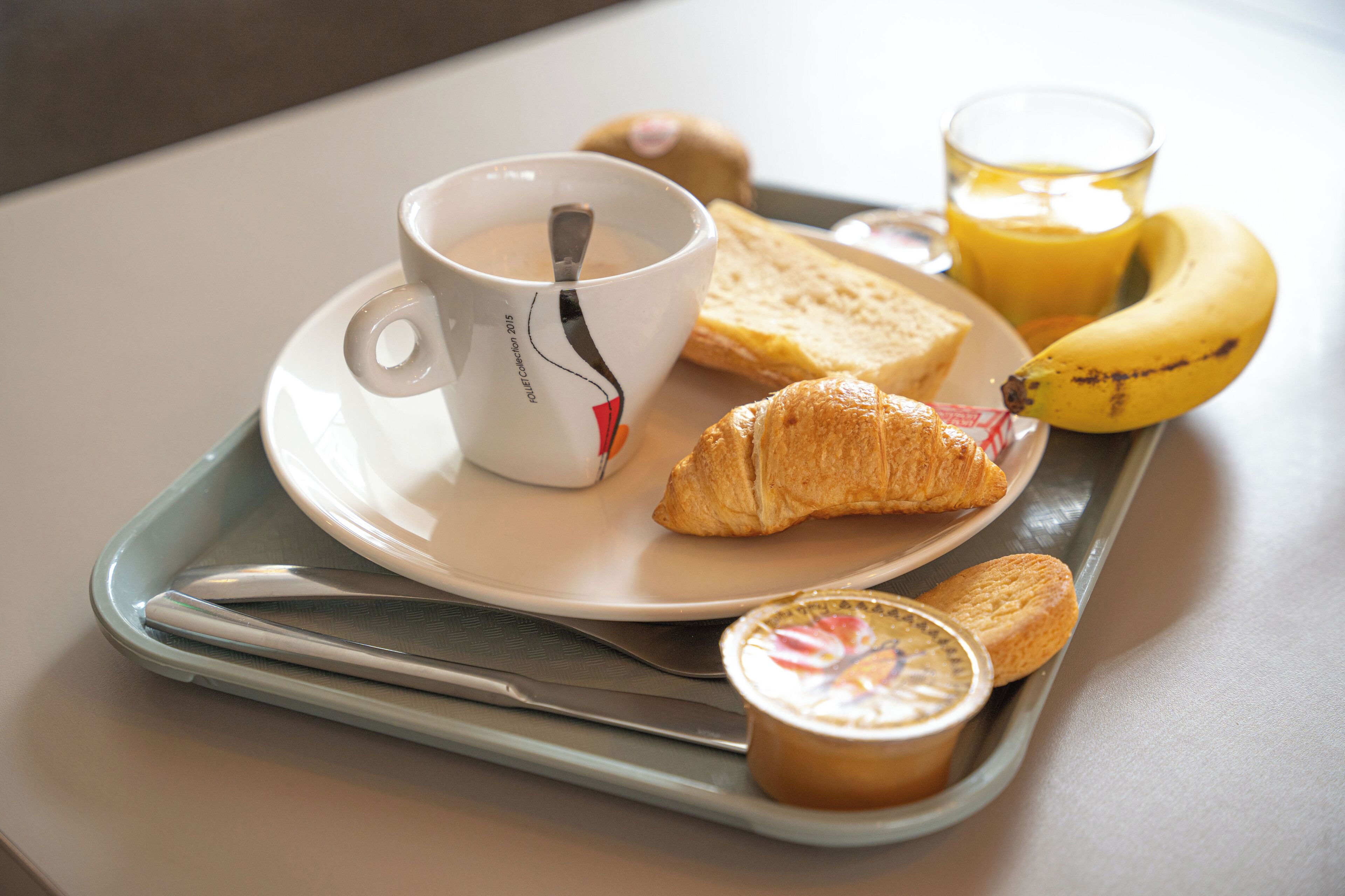 Ein Tablett mit einer Kaffeetasse, einem Croissant, einer Banane, einem Glas Orangensaft und einer kleinen Joghurtportion.