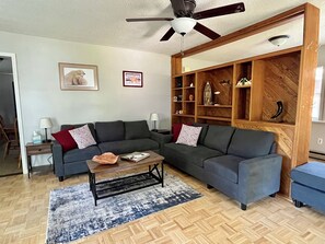 A living room with a dark grey sectional sofa, a coffee table, a patterned rug, a wooden shelving unit, and a ceiling fan.