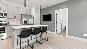 A modern kitchen with a central island, bar stools, a flat-screen TV, and a grey wall. - The *Minimalist's Escape* near Greenbelt & Winery with HOT TUB! (Garden City)