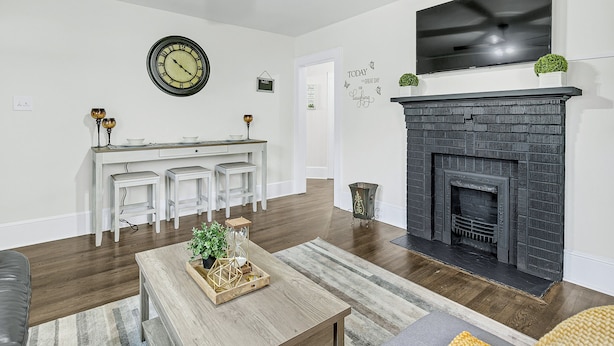 Una sala de estar con chimenea, un televisor montado encima, un reloj en la pared y una zona de cocina con armarios blancos y taburetes.