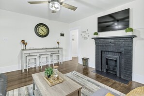 Una sala de estar con chimenea, un televisor montado encima, un reloj en la pared y una zona de cocina con armarios blancos y taburetes.
