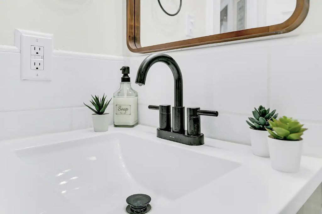 A white bathroom sink with a black faucet, a soap dispenser, and two small potted plants.