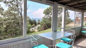 A covered patio with a table and chairs, large windows offering a view of trees and a distant landscape. - Private Beach.  Fabulous Bay Views. WIFI (Brewster)