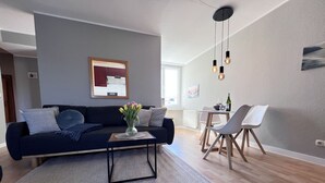 A modern living room with a dark blue sofa, a coffee table, and a dining area with a white table and chairs. - Fewo Strandgut - "Strandgut" ... close to the beach and cozy! (Dahme)