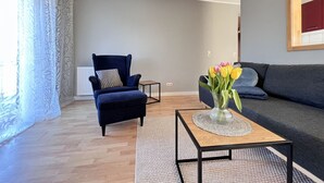 A living room with a dark blue armchair, a grey sofa, a small wooden coffee table with a vase of flowers, and patterned curtains. - Fewo Strandgut - "Strandgut" ... close to the beach and cozy! (Dahme)