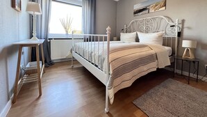 A bedroom with a bed, bedside tables, a window with curtains, a radiator, and a painting on the wall. - Fewo Strandgut - "Strandgut" ... close to the beach and cozy! (Dahme)